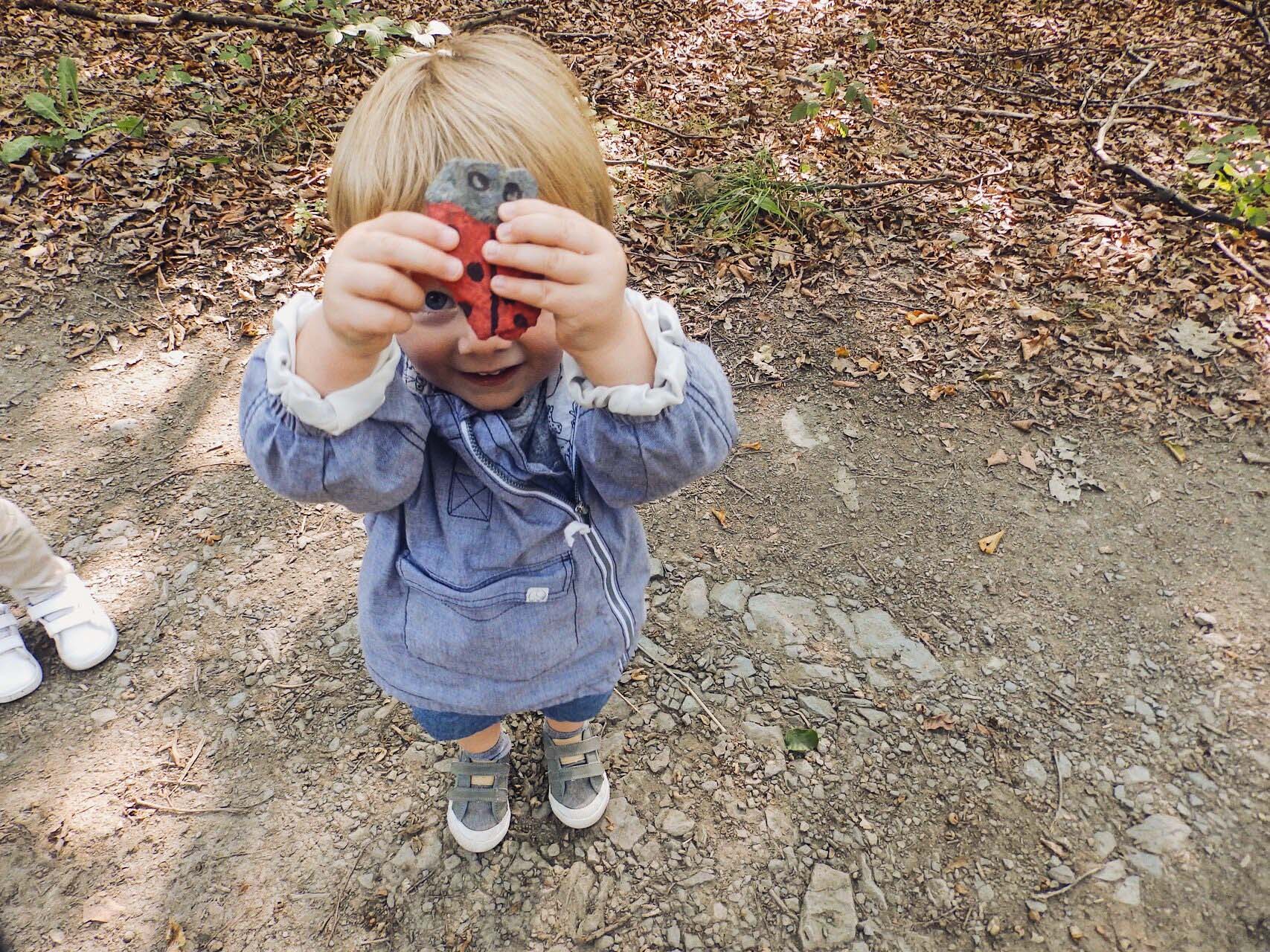 Un enfant tient en l'air une coccinelle qu'il a bricolée, de manière à cacher son visage. Il est dehors et a un sourire éclatant. Nous essayons d'organiser de nombreuses activités à l'extérieur afin de stimuler l'esprit de découverte des enfants, de renforcer leurs défenses immunitaires et de construire une relation positive avec la nature et l'environnement.