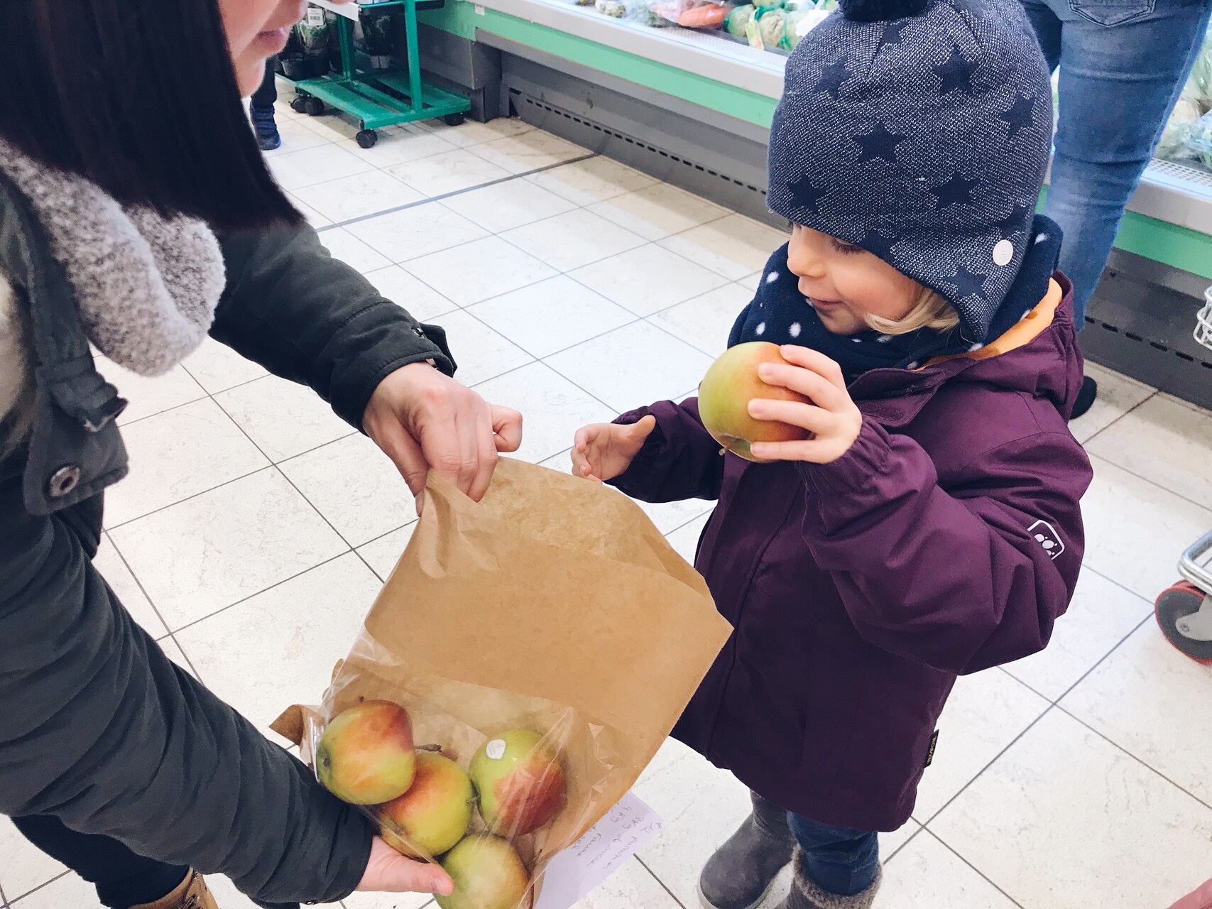 Une femme tient quelques pommes dans un morceau de papier et les offre à un enfant. L'enfant tient une pomme dans sa main mais regarde l'autre pour voir s'il a fait le bon choix. Pour Butzemillen Crèches Luxembourg, il est important que les enfants reçoivent une alimentation équilibrée et saine dès leur plus jeune âge.
