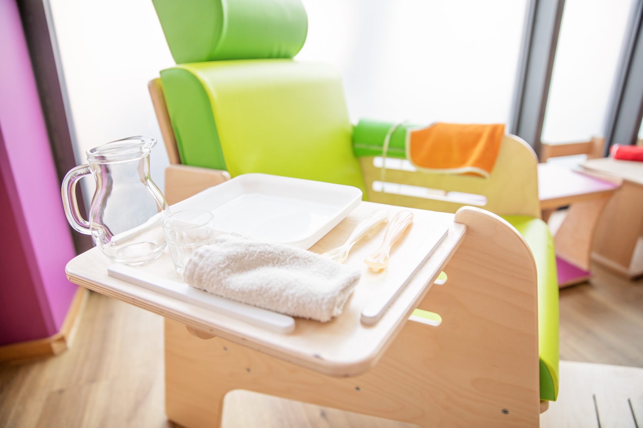 Sur l'image, on voit une chaise Pikler. Elle est en bois et un coussin est posé dessus. La planche qui sert à manger est tirée vers l'extérieur. Sur cette planche se trouvent une assiette, une carafe et des couverts. Dans nos crèches, nous veillons à ce que les enfants puissent avancer dans la vie de manière autonome dès leur plus jeune âge. C'est pourquoi nous utilisons la méthode Pikler, dans laquelle l'espace joue un rôle essentiel. Lors des quatre repas quotidiens, nous veillons à ce que les plats soient équilibrés et sains.