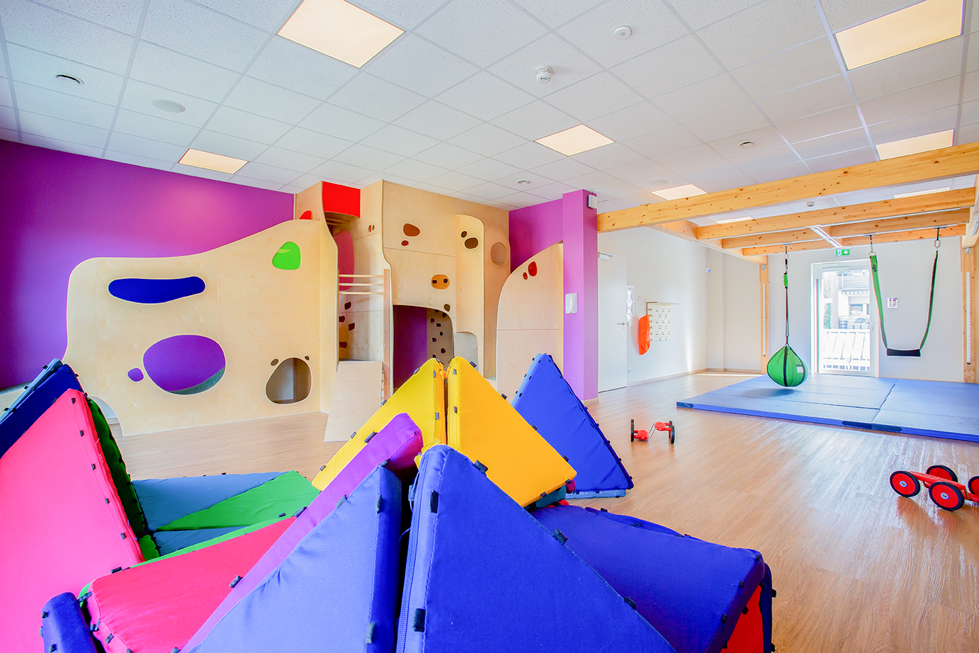Une autre photo d'un intérieur coloré montre la diversité de l'équipement. On y voit un mur d'escalade, des jouets modulaires, une balançoire et divers autres jouets. La diversité des jouets encourage la créativité et le développement individuel.