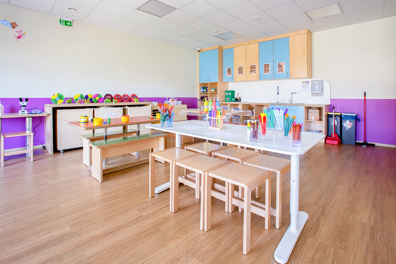 Au premier plan se trouve un grand bureau avec 4 places assises de chaque côté. On y trouve des crayons, des ciseaux et d'autres fournitures. Ces tables communes sont utilisées pour le bricolage, l'apprentissage, la peinture et d'autres activités. En apprenant ensemble, on peut apprendre les uns des autres et, en travaillant de manière créative en compagnie, on s'inspire mutuellement.