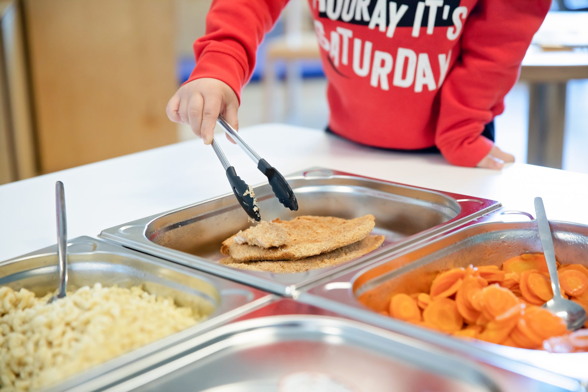 Un enfant se tient devant un buffet. DIverse récipients en acier sont ouverts on voit une escalope, des carottes et du riz complet. L'enfant est en train de prendre une escalope avec une pince métallique. Il est important pour nous que les enfants de nos crèches composent eux-mêmes leurs repas et apprennent ainsi dès leur plus jeune âge ce que signifie une alimentation saine.