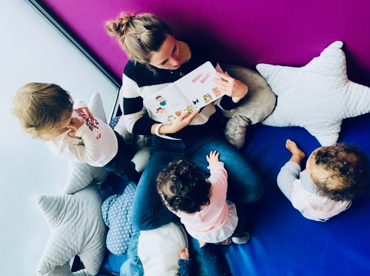 Une femme est assise sur le sol et montre un livre à trois enfants qui regardent le livre dans la cour. Sur le sol, on distingue quelques coussins en forme d'étoile. Grâce à de telles activités communes, les enfants apprennent les uns des autres et la dynamique de groupe, ainsi que les compétences sociales de chacun, sont encouragées.