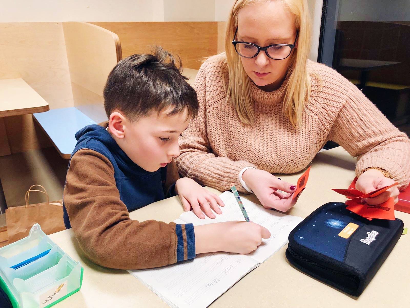 Une femme est assise à côté d'un enfant qui est penché sur un cahier. Les devoirs de nos crèches préparent les enfants à la rentrée scolaire. En donnant aux enfants des devoirs à domicile, nos crèches impliquent les parents dans l'éducation et nous aident à préparer les enfants à leur scolarité. D'autre part, les enfants ont ainsi la possibilité de partager leur expérience avec leurs parents.
