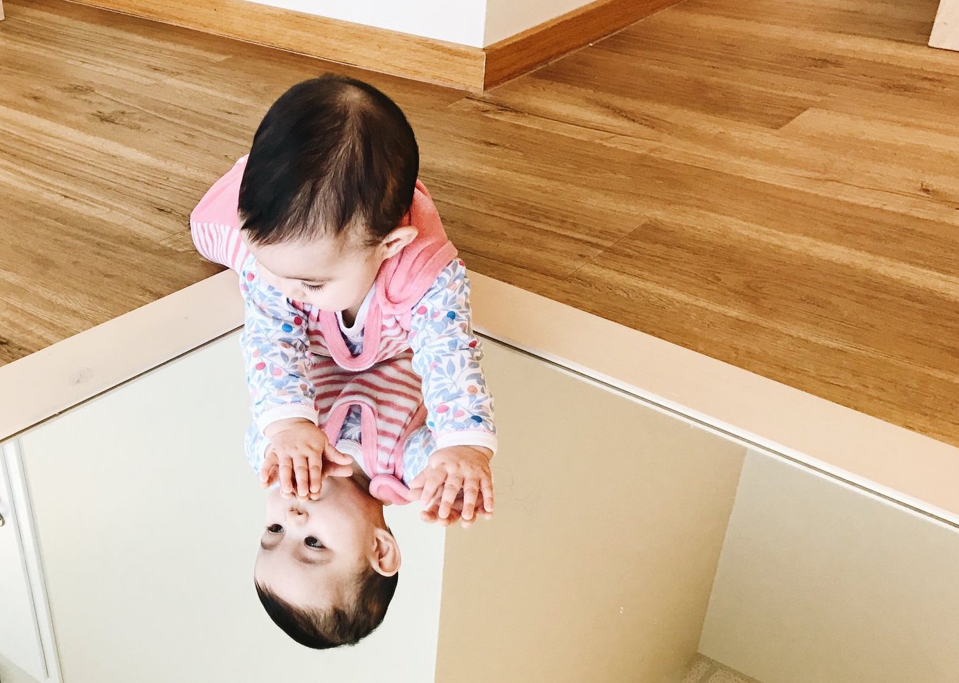 Un enfant habillé de couleurs vives est allongé sur le sol et se regarde dans un miroir. Il se regarde, l'air pensif. Avec cette image, nous voulons souligner que dans nos crèches, les enfants ont la possibilité de réfléchir sur eux-mêmes dès leur plus jeune âge.