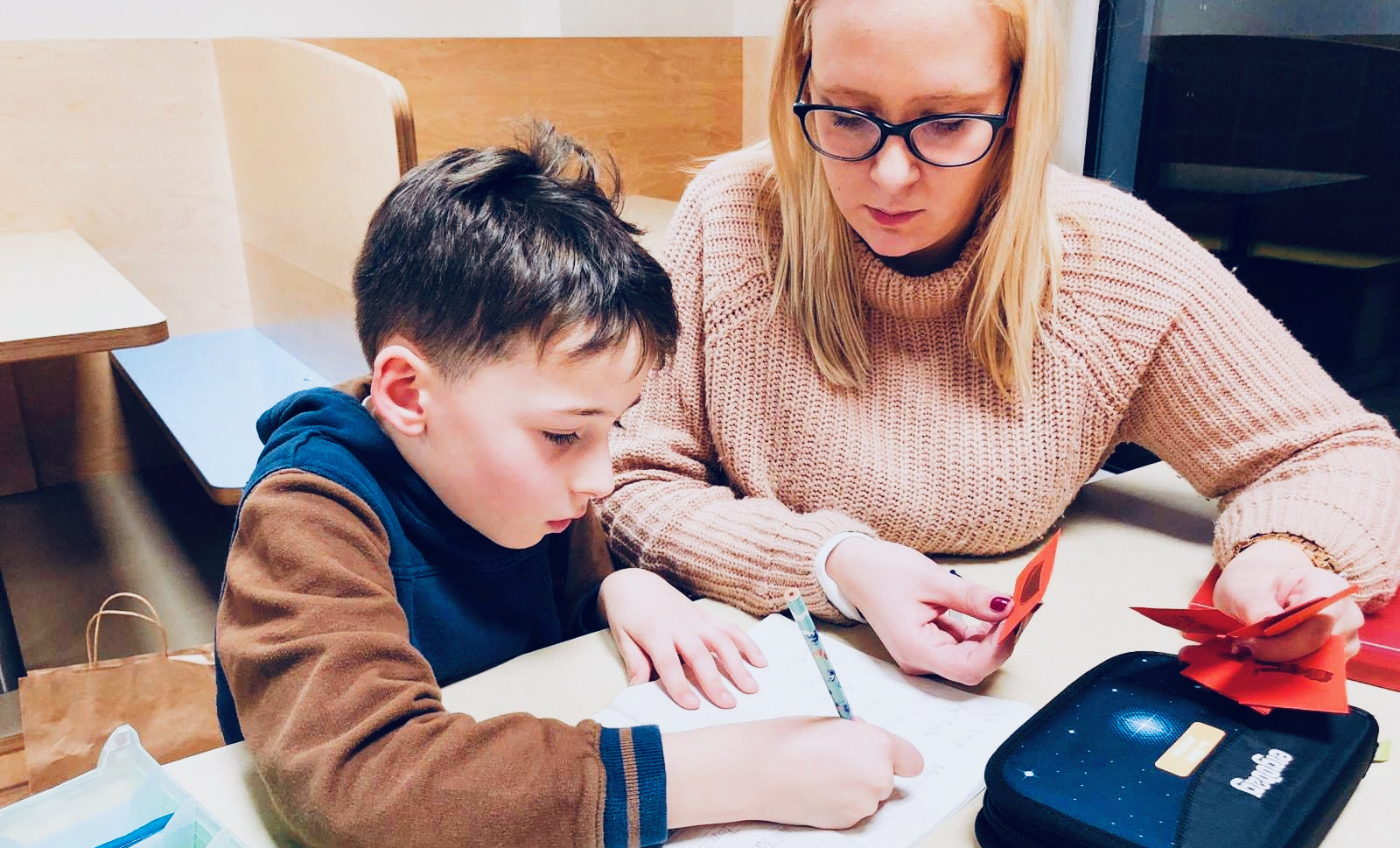 Un jeune est assis avec une monitrice pour faire ses devoirs. L'aide aux devoirs fait partie du quotidien des crèches. Dans une atmosphère calme, la concentration est favorisée et comme les devoirs ont déjà été faits à la crèche, les familles peuvent se concentrer sur leur vie de famille après le travail.