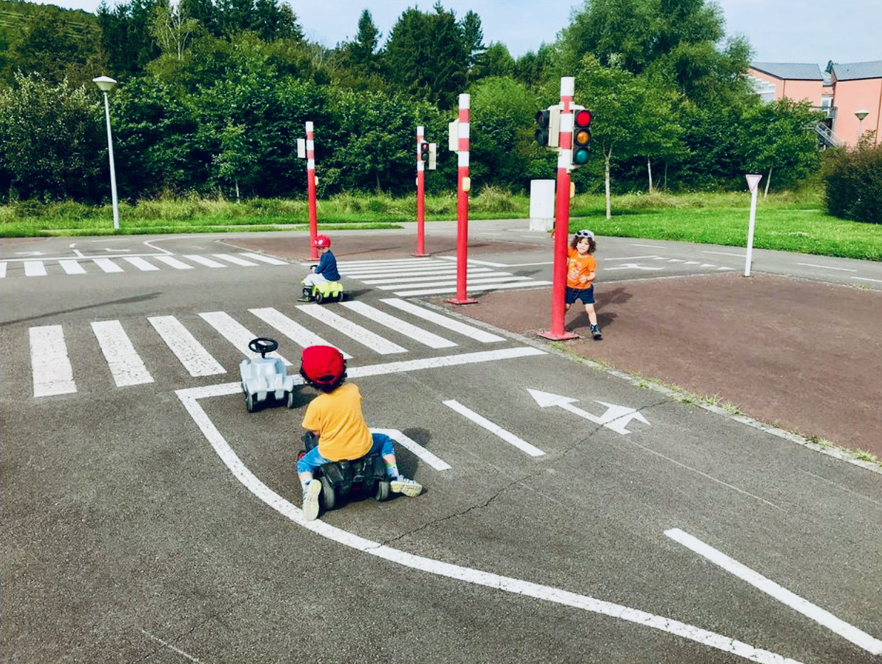 On voit le parcours routier d'une de nos crèches qui est prévu pour les bobbycars. A un carrefour, un enfant est debout sur sa voiture devant un feu rouge, devant lui se trouve un véhicule vide. Au feu rouge, un enfant attend en tant que piéton. De telles infrastructures aident les enfants à s'expliquer le monde qui les entoure, favorisent les déductions logiques et la compréhension des relations entre les différents facteurs de leur environnement.