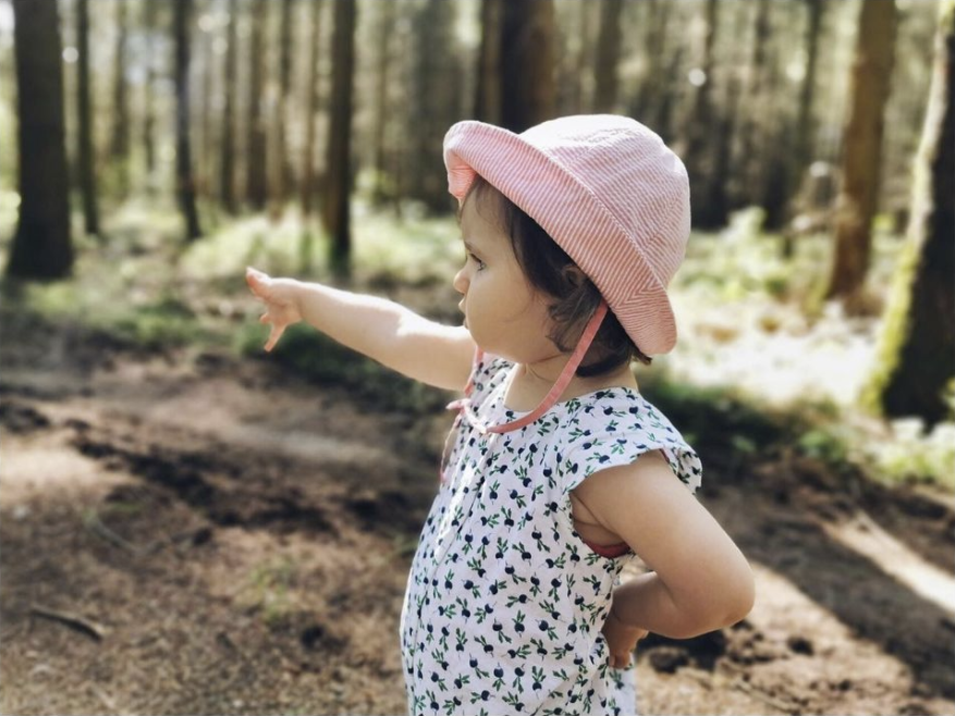 Une jeune fille se trouve dans la forêt, les bras écartés. Elle porte un chapeau rose et un t-shirt à motifs bleus et blancs. Elle se trouve dans la partie inférieure droite de l'image et ses bras écartés, ainsi que son regard, sont dirigés vers la partie supérieure droite de l'image. L'image symbolise la proximité avec la nature qui est donnée par la situation de nos sites.